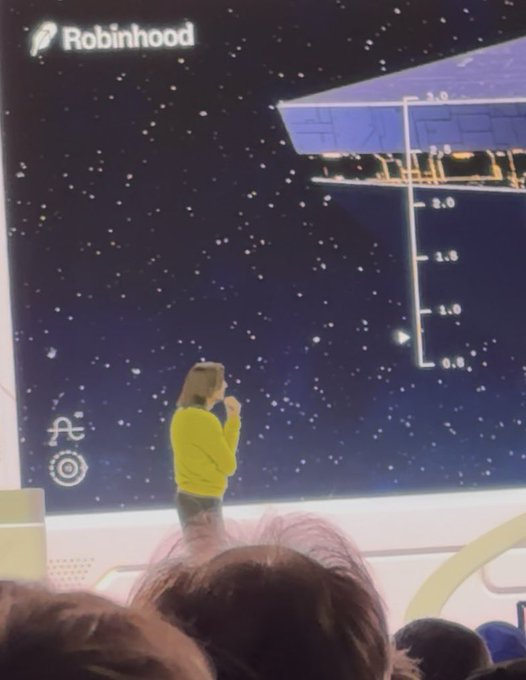 A photo of a presentation stage featuring a woman in a yellow sweater speaking in front of a starry space-themed backdrop displaying the Robinhood logo and various interface elements.