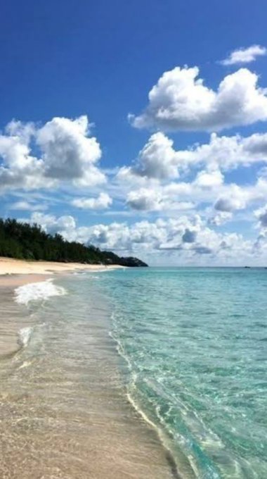 A tropical beach scene with turquoise water, gentle waves, and a blue sky filled with fluffy white clouds.