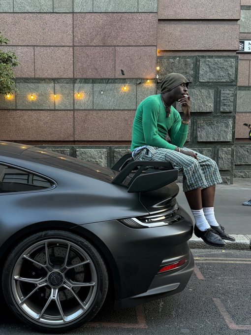 Person wearing a green sweater, plaid pants, and a beanie sitting on the rear spoiler of a matte black Porsche sports car on a city street.