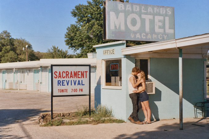 A couple embraces in front of a turquoise motel office with a large "BADLANDS MOTEL VACANCY" sign overhead, while a sign advertises a "SACRAMENT REVIVAL TONITE 7:00 PM."