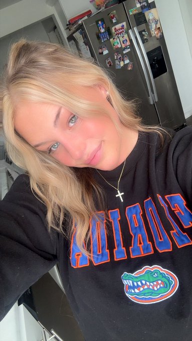 A blonde woman smiles in a selfie, wearing a black Florida Gators sweatshirt and a cross necklace, with a refrigerator covered in magnets visible behind her.