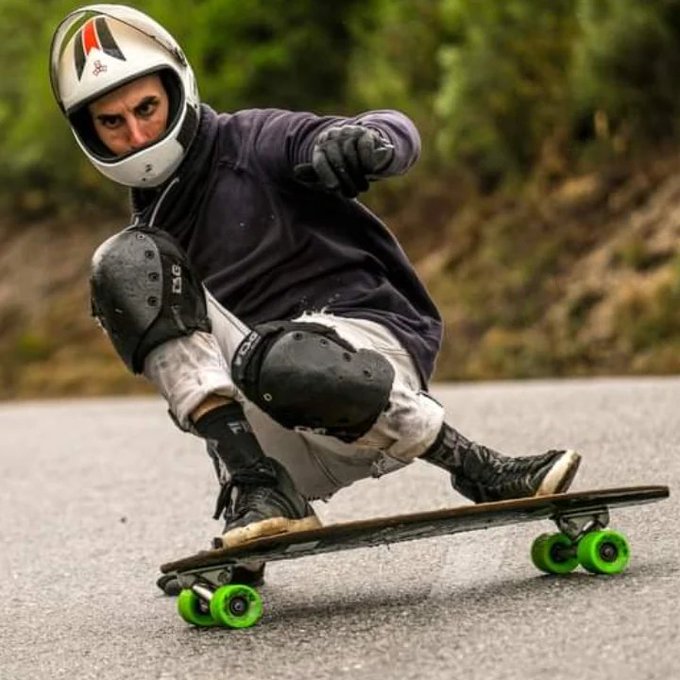 A skateboarder wearing a full-face helmet and knee pads rides a longboard downhill, crouched low with one hand reaching toward the pavement.