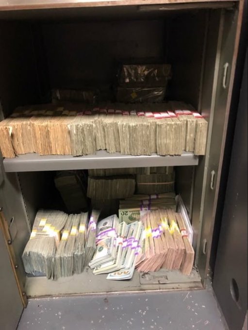 An open safe filled with shelves of banded stacks of cash, including visible $20 bills, alongside wrapped packages on the top shelf.