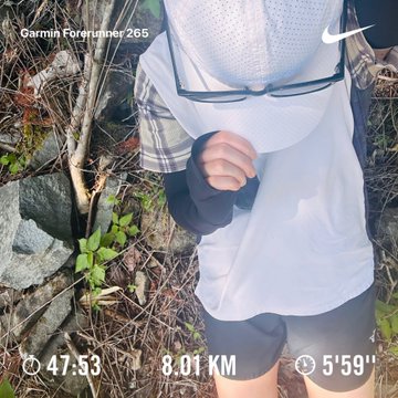 The image features a person wearing shorts, a white shirt, and glasses while standing in a field or forest area. The person appears to be taking a break from their outdoor activities, as they are holding up a pair of sunglasses for the camera. The background consists of trees and rocks, creating an impression of being outdoors on a sunny day.