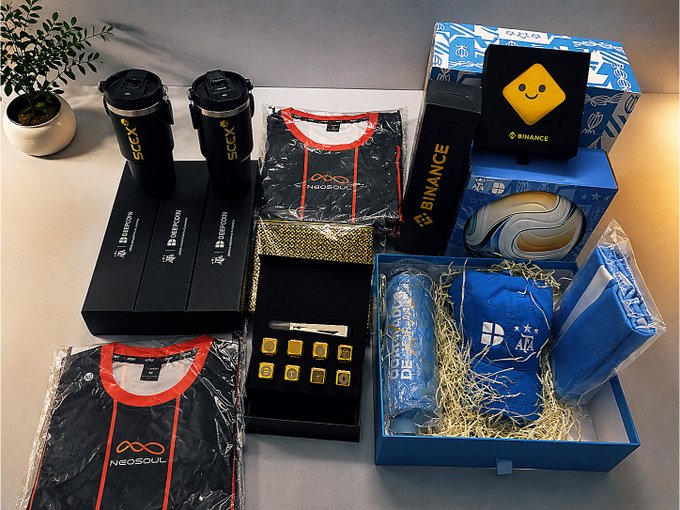 A collection of branded merchandise including Binance water bottles, NeonSoul t-shirts, a Bitcoin-branded tumbler, a blue baseball cap, and gold bars in black presentation boxes, along with various packaged items.