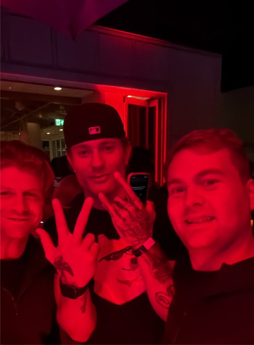 Three men pose for a selfie in a room with red lighting; one wears a black baseball cap and makes a peace sign with tattooed hands.