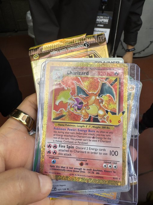 A hand wearing a gold ring holds a holographic Charizard Pokémon card (120 HP, Fire Spin attack) alongside a Pikachu promo card.
