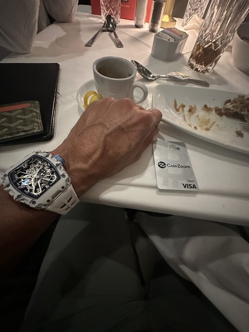 A person's wrist wearing a white Richard Mille watch while seated at a restaurant table with a CoinZoom debit Visa card, coffee cup, and empty plate.