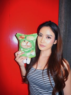 A young woman with long dark hair wearing a striped top poses against a red background, holding up a green package of snacks.