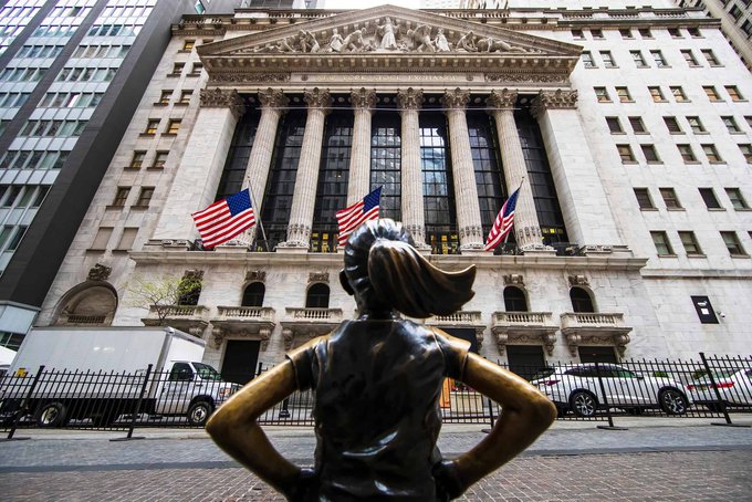 The "Fearless Girl" statue stands defiantly in front of the New York Stock Exchange, where American flags fly alongside the building's iconic neoclassical columns. This powerful image of female resolve at the heart of Wall Street symbolizes the traditional financial establishment, contrasting with the post's message about the entire stock market transitioning onto blockchain technology.