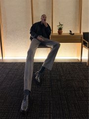 A man sits on a wooden bench in a modern, warmly lit room with potted plants, looking off to the side. The accompanying post teases $ANSEM as "the next leg's gonna be a big one," suggesting the man pictured may be the trader/influencer Ansem himself, using the casual candid photo to build hype around the ticker.