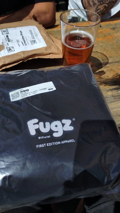 A "Fugz Official First Edition Apparel" package sits on a wooden table next to a glass of amber beverage, with another similar delivery package visible in the background, capturing the unboxing moment at a collectors' meetup.