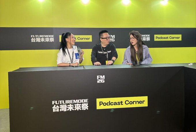 Three people sit at a black desk in a podcast studio with yellow walls, featuring 'FUTUREMODE Podcast Corner' and 'FM 26' signage on the desk and backdrop.