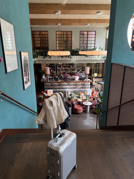 A silver suitcase draped with a tan coat stands at the top of a staircase overlooking a stylish bar lounge with teal walls and large pendant lights. The scene captures a moment of arrival in Belgium, where the traveler celebrates KuCoin's MiCa license at the Tomorrowland festival.