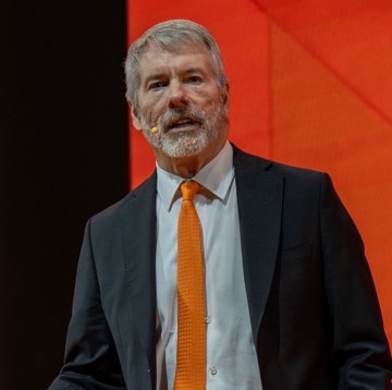 A man with a beard, wearing a dark suit, white shirt, and bright orange tie, speaks on stage against a red backdrop, likely delivering a keynote about Strategy's dramatic $22.7B unrealized swing on its Bitcoin holdings.