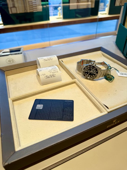 The image shows a display case with two watches and a credit card placed inside of it. The watches are positioned next to each other, one being a Rolex watch and the other a wristwatch. A black wallet can also be seen on the display case. The credit card is placed in front of the Rolex watch.