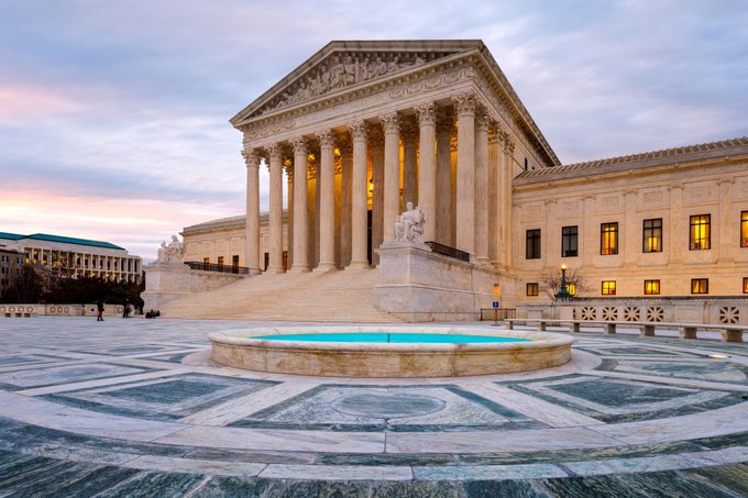 The U.S. Supreme Court building stands majestically at dusk, its grand neoclassical columns illuminated against a pastel sky, with a circular fountain reflecting in the foreground plaza. The image serves as a fitting visual metaphor for the legal and regulatory hurdles facing the CLARITY Act, as eight banking groups push for tighter stablecoin rules ahead of today's Senate vote.