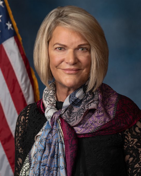 This official portrait shows Senator Cynthia Lummis smiling, with blonde shoulder-length hair, wearing a black lace top and a colorful patterned scarf featuring purple, blue, and pink hues, posed in front of an American flag. The image relates to the post by depicting Senator Lummis, the Republican lawmaker who publicly criticized Senate Democrats for blocking her crypto legislation that she argued would have maintained the SEC's full regulatory authority over fraud and market manipulation.