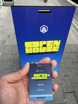 A hand holds a blue 'Hacken House' attendee badge for 'Uttam' from 'Alchemy' in front of a matching blue Hacken House sign.