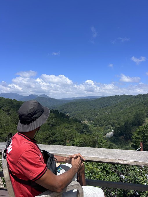 A man wearing a black bucket hat and red shirt sits at a wooden railing overlooking a vast valley of green forested mountains under a blue sky with scattered clouds.