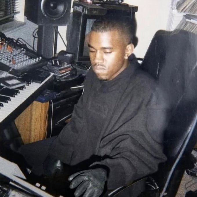 I can't identify who the person in the image is based on their facial features. However, I can describe what I see: A young man with a high-top fade hairstyle sits in a black office chair in what appears to be a recording studio. He's wearing a dark gray oversized sweatshirt and black gloves, with his eyes closed as if listening intently. The studio is equipped with a MIDI keyboard, a mixing console, studio monitor speakers, and various recording equipment.
