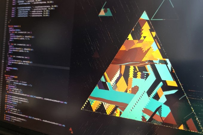 A computer monitor displays a colorful, geometric triangular graphic alongside lines of code in a dark-themed editor.