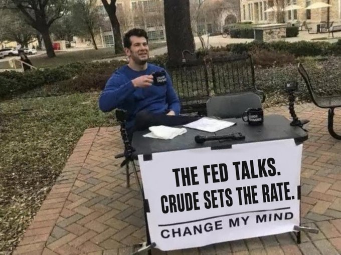 Man at a 'Change My Mind' table with a sign reading 'THE FED TALKS. CRUDE SETS THE RATE.' holding a coffee mug outdoors.