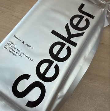 A glossy silver "Seeker" phone box by Solana Mobile rests on a wooden surface, displaying the bold black "seeker" branding along with text reading "SOLANA 📱 MOBILE" and "GENESIS PHONES AND DISTRIBUTED BY SOLANA MOBILE." The packaging directly ties to the post's excitement about finally purchasing the Seeker device after watching others engage with the Solana mobile ecosystem.