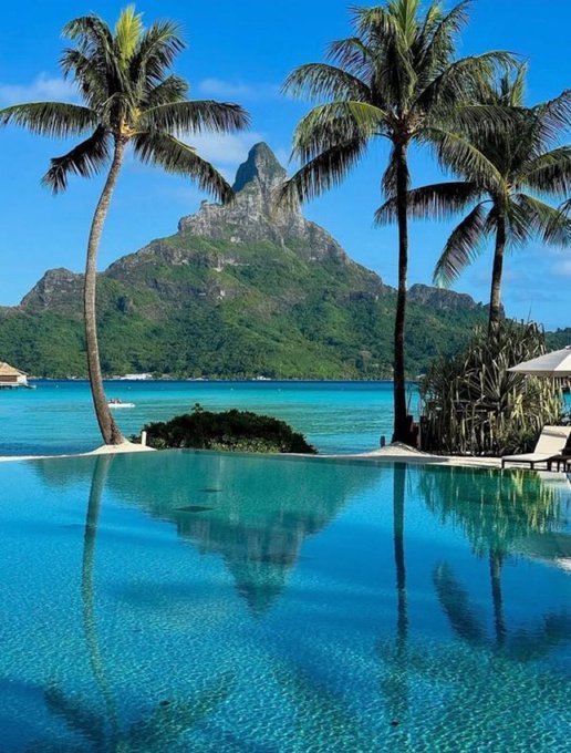 A stunning infinity pool reflects a tropical paradise scene, with palm trees framing a majestic mountain rising from crystal-clear turquoise waters. The serene, vacation-like imagery mirrors the post's upbeat mid-week promotion for RiverdotInc's chain-abstraction stablecoin system, evoking the same relaxing, "stay-tuned" vibe as their ongoing River S6 campaign.