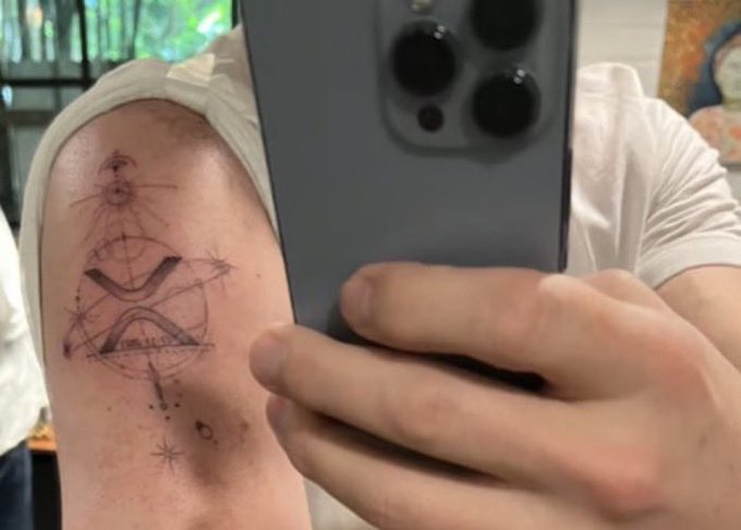 A mirror selfie showing a person's upper arm with a tattoo featuring an eye, geometric shapes, and an 'X', partially obscured by a smartphone with a triple camera.