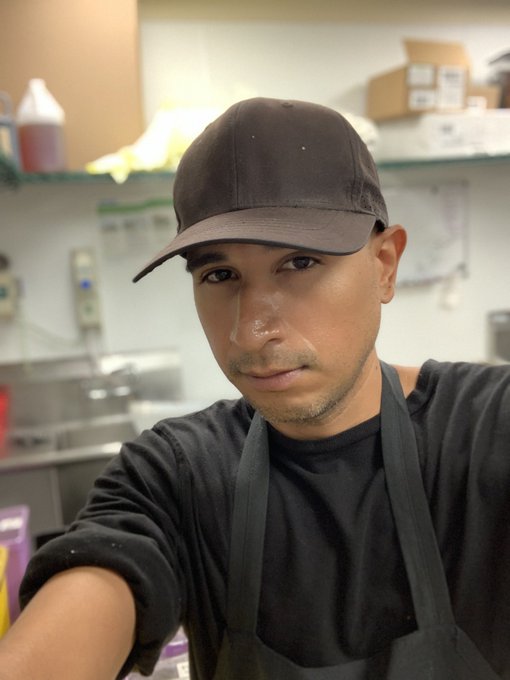 A selfie of a young man wearing a black cap and apron, seemingly taken in a restaurant or kitchen setting.