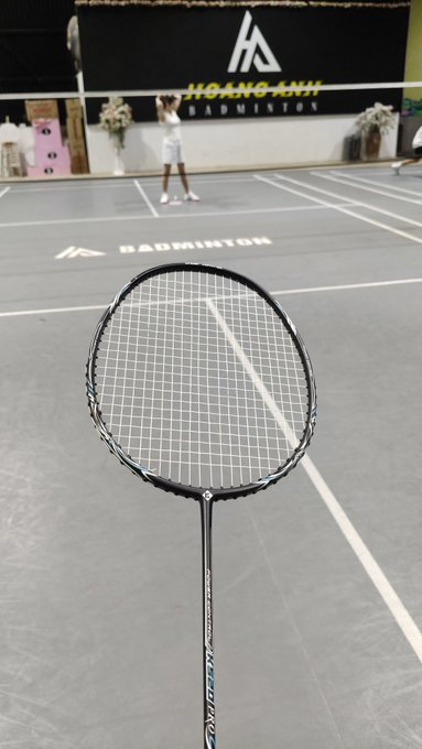 A first-person view of a badminton racket on a gray court at 'HOANG ANH BADMINTON' facility, with a player visible across the net.