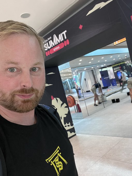 A selfie of a red-haired, bearded man in a black t-shirt standing in front of an entrance to the 'SOLO SUMMIT BERLIN' event, with an inflatable sports court visible inside.