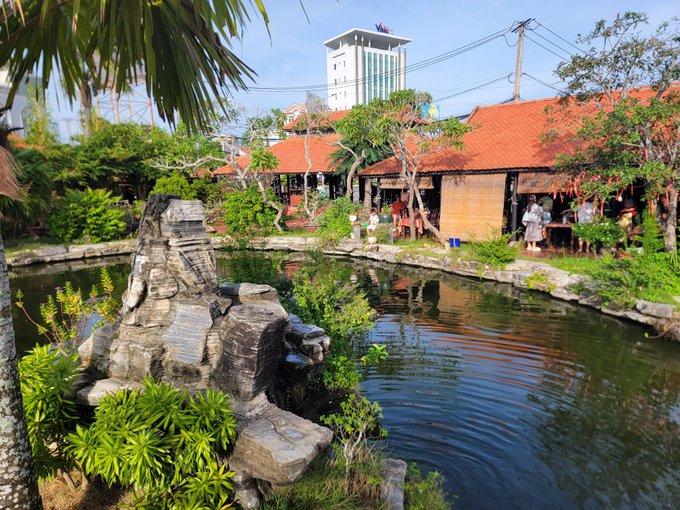 A scenic view of a pond with koi fish surrounded by tropical vegetation and traditional buildings with red tile roofs, with a tall white building in the background.