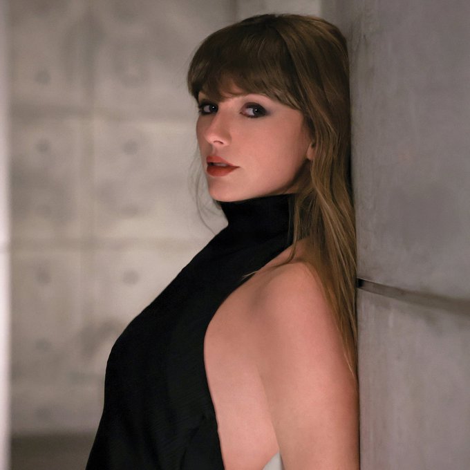 A portrait of a woman with long brown hair and bangs, wearing a black halter top, leaning against a gray textured wall.