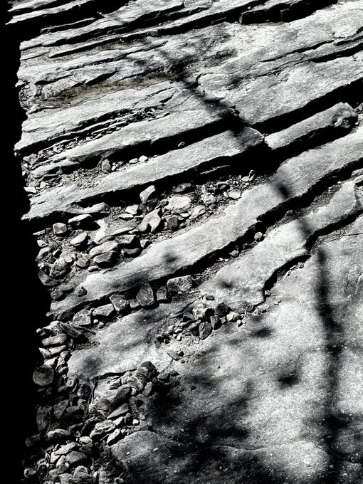 A black and white photograph of layered, cracked stone slabs and rock fragments, possibly ancient ruins or geological formations, with shadow patterns across the surface.
