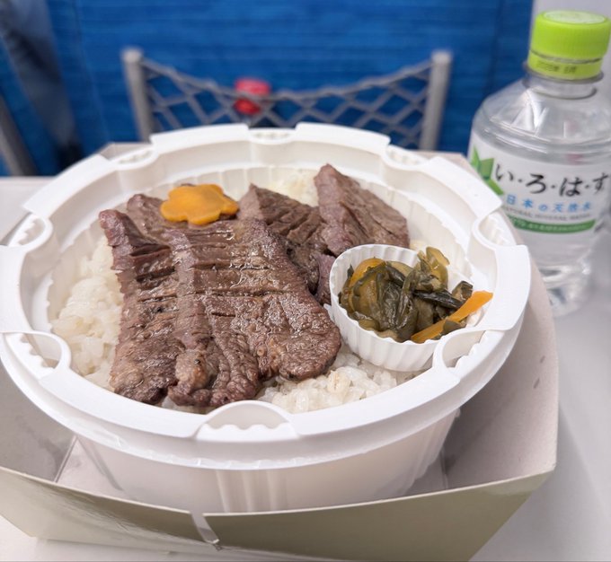 A takeout bowl featuring sliced grilled beef over white rice, accompanied by a small side of pickled vegetables in a flower-shaped dish, next to a bottle of Japanese mineral water with a green cap and label reading 'いろ・はす' (Irohasu).