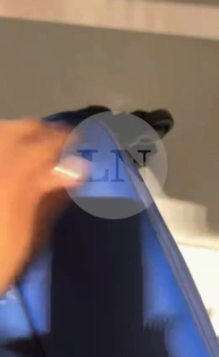 The image shows a person's hand holding a blue object, possibly a piece of paper or a book, with their thumb on top and fingers below. The background appears to be a bathroom setting, as indicated by the presence of a sink and a mirror in the scene.