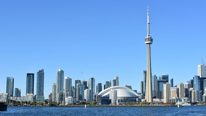 A clear daytime photograph of the Toronto skyline featuring the CN Tower, the Rogers Centre dome, and Lake Ontario in the foreground.