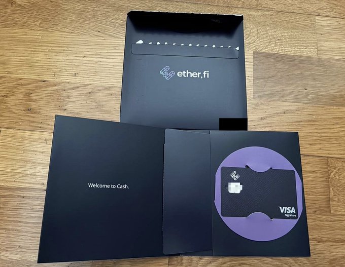 A black ether.fi/Visa debit card and its packaging are displayed on a wooden floor, with the card's envelope reading "Welcome to Cash." The card holder has just received this physical version of their crypto-backed card after reaching the Luxe tier, having previously only used the virtual one.