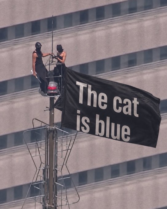 Two figures dressed in black are perched atop a tall metal mast or antenna structure, having unfurled a large black banner that reads "The cat is blue" in white lettering, with a multi-story building visible in the background. The banner and the caption "They know." together create an eerie, conspiratorial tone, suggesting that the activists have revealed some hidden truth or secret message directed at an unknowing audience below.