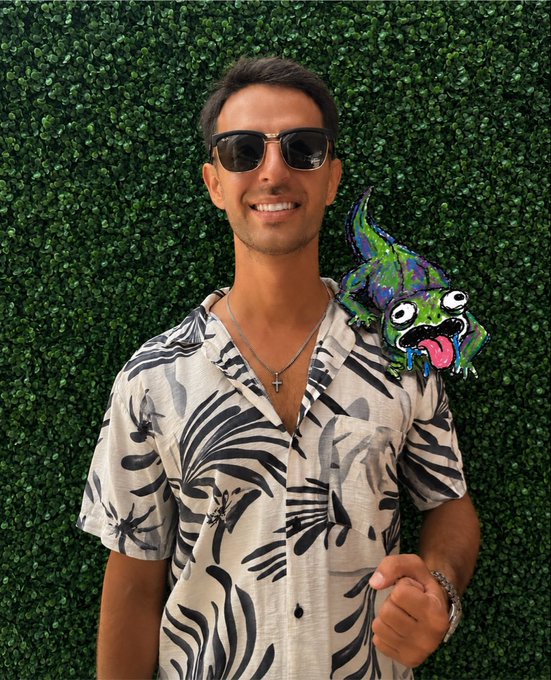 A man in sunglasses and a tropical-print shirt smiles against a green hedge backdrop, with a colorful cartoon chameleon illustration on his shoulder. The playful image complements his post by using the "still here" chameleon reference to symbolize resilience in the crypto and NFT space while teasing his upcoming project.