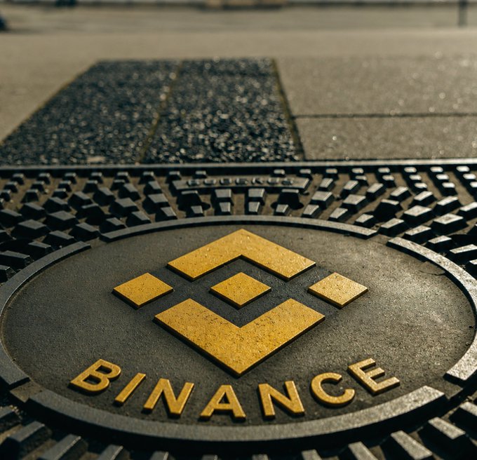 A metallic Binance manhole cover sits flush with a city sidewalk, featuring the platform's gold logo and "BINANCE" name in bold lettering—fitting the "streets are talking" caption as the crypto exchange literally marks its presence on the streets.