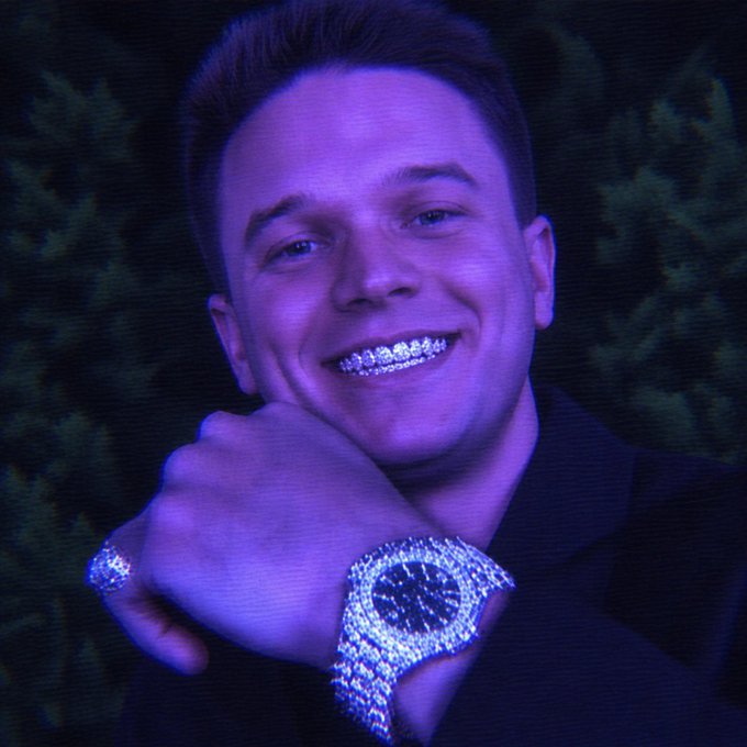 A young man smiles broadly at the camera, flashing diamond-encrusted grillz on his teeth along with a flashy jeweled watch and matching ring, all bathed in a moody purple light. The opulent, jewelry-heavy look visually mirrors the post's hype around Stake's monthly giveaway and "free money" theme.