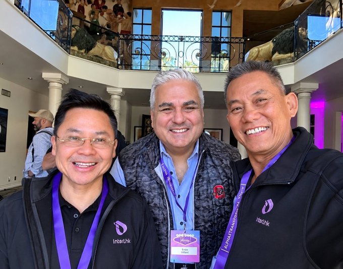 Three smiling men pose for a selfie at the Alpha Transform Conference, with two wearing black 'Interthnk' jackets and purple lanyards, standing in a brightly lit atrium with balconies.