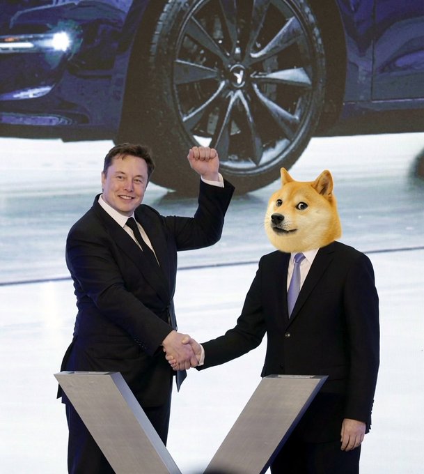 The image features two men dressed in suits, with one man wearing a dogecoin mask that reads "Elon Musk will become the first Dogecoin Trillionaire in history tomorrow." The other man has his fist raised in a handshake. The background shows an advertisement for a car and a wheel.