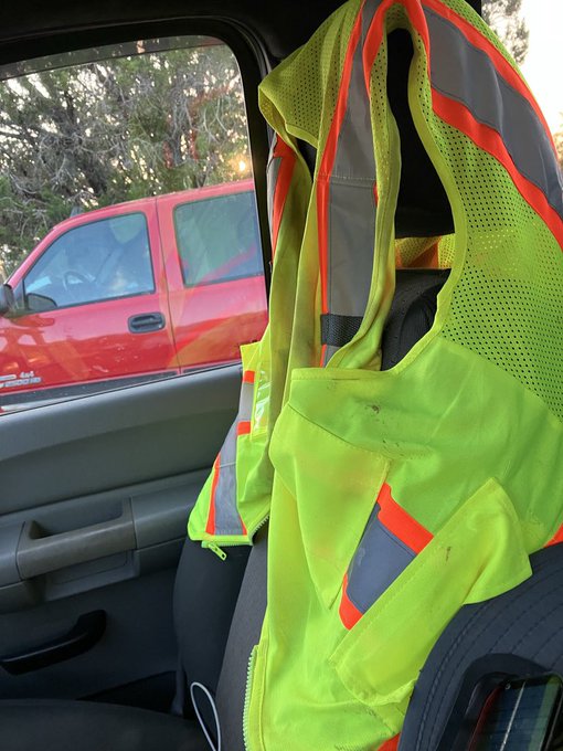 A high-visibility yellow safety vest with orange reflective stripes hangs inside a vehicle, with a red pickup truck visible through the window.