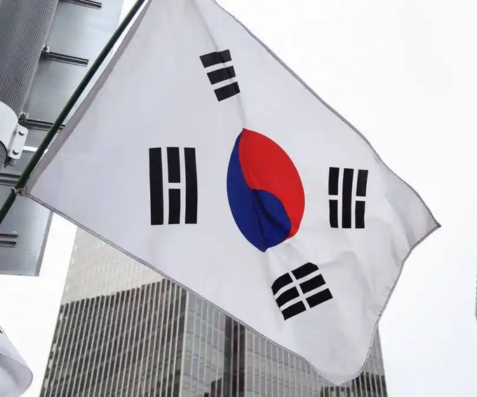 The South Korean national flag (Taegukgi) waving against an overcast sky, with a high-rise building visible in the background.