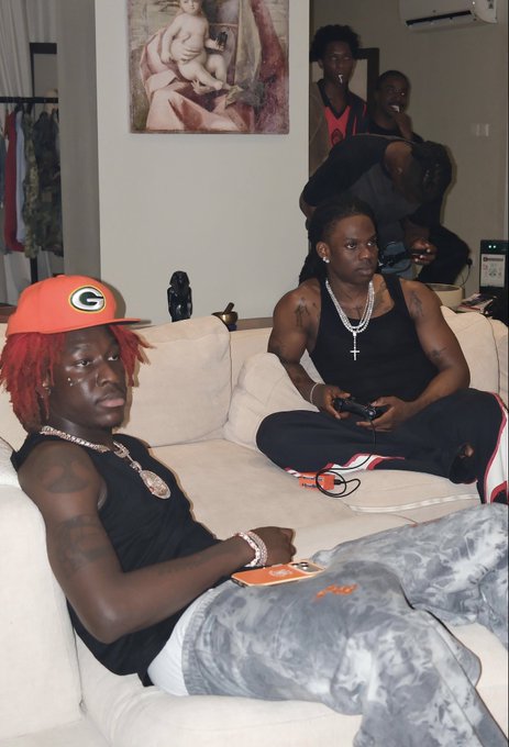 Two men relax on a beige couch in a living room; one wears an orange Green Bay Packers cap with red dreadlocks, while the other, in a black tank top with cross necklaces, plays a video game.