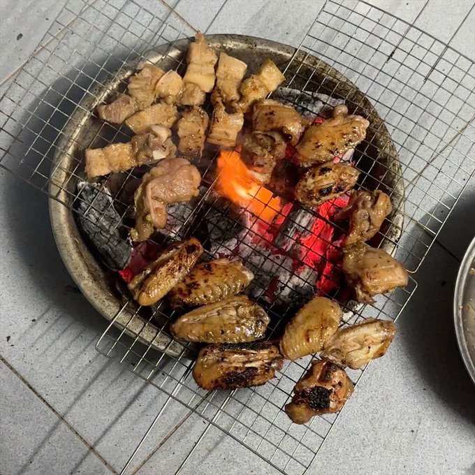 Pieces of meat and pork belly grilling on a wire mesh over a charcoal brazier with visible flames.